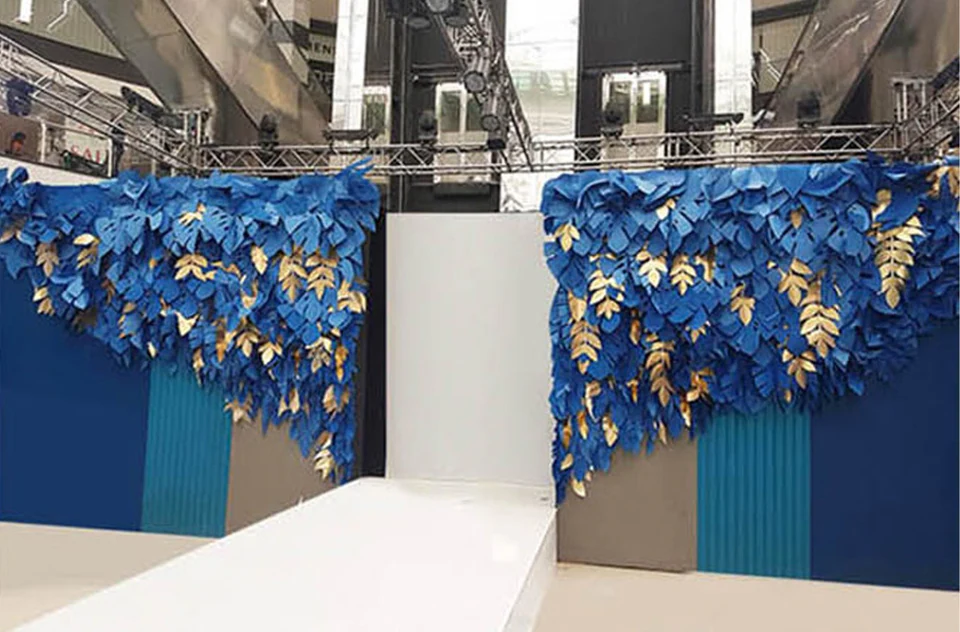 A runway framed by walls of blue paper monstera leaves and gold leaves under a lighting rig