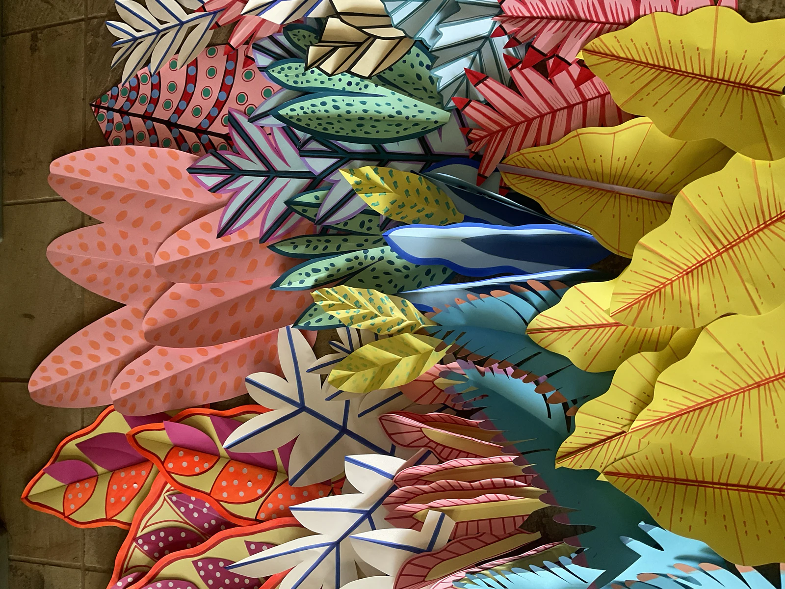 Hand-painted paper leaves with spots, stripes and veins, laid out on a tiled floor, seen from above